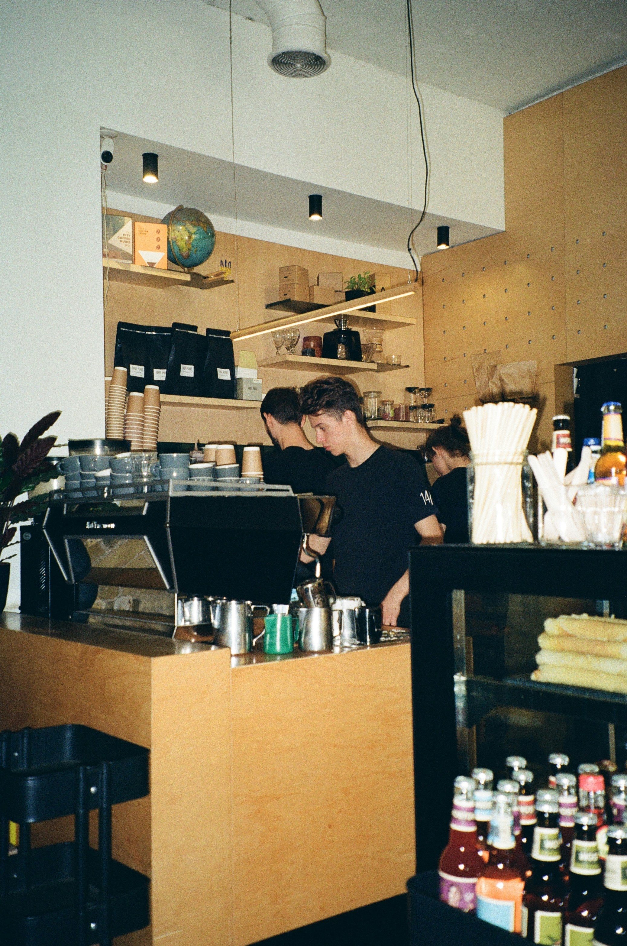 A warm, slightly grainy interior shot of a cafe or coffee shop counter, showing baristas working behind a wooden espresso machine and counter, with shelves and bottles in the background.